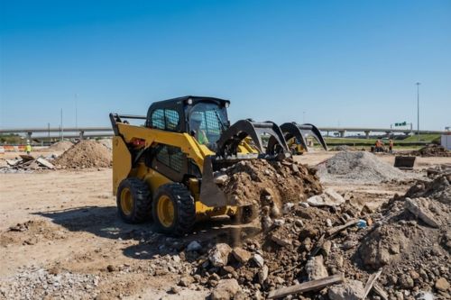 Operator in a yellow skid steer loader with a grapple attachment clearing dirt and construction debris at an active Houston-area job site.